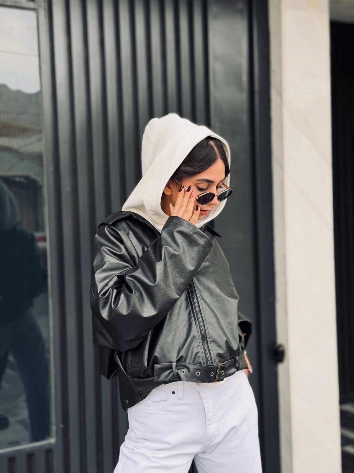 Woman in black leather jacket, white hoodie, and white jeans wearing sunglasses.