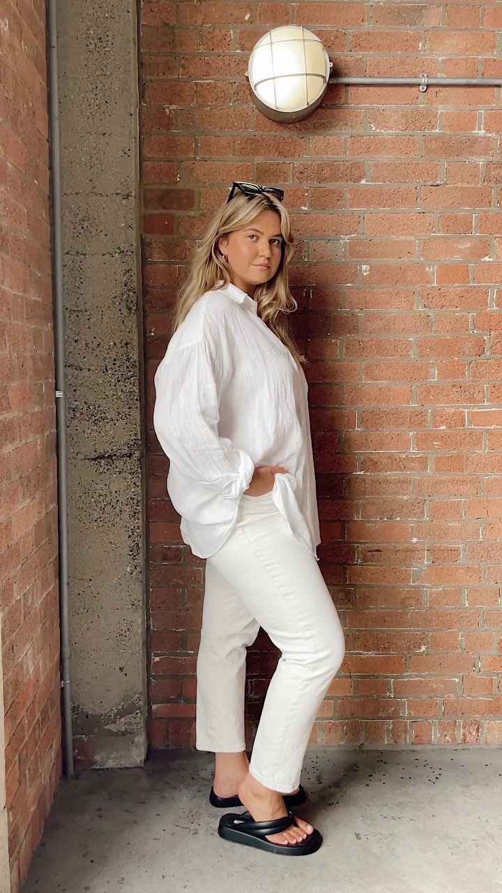 Woman wearing an oversized white linen shirt and slim white jeans against brick wall