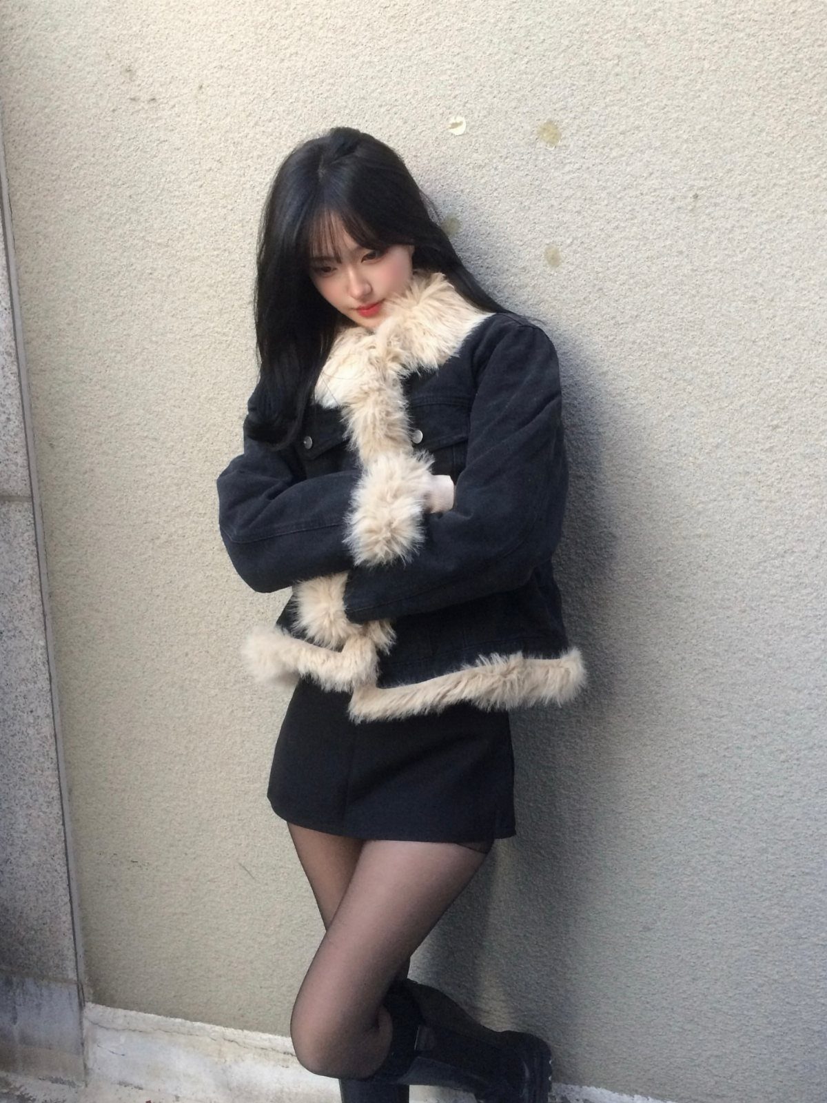 Woman in a black faux-fur trimmed jacket and mini skirt posing against a wall.