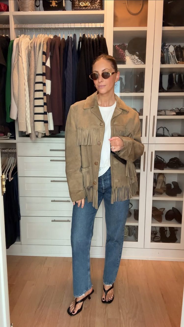 Woman wearing a tan suede fringe jacket, white shirt, and blue jeans in a closet.