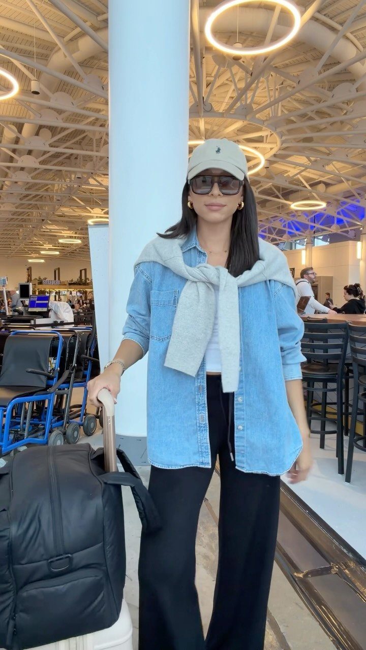 Women Travel Outfit: 8 Looks for Airport to City 2 Woman in denim shirt and wide-leg pants posing with luggage in an airport terminal.