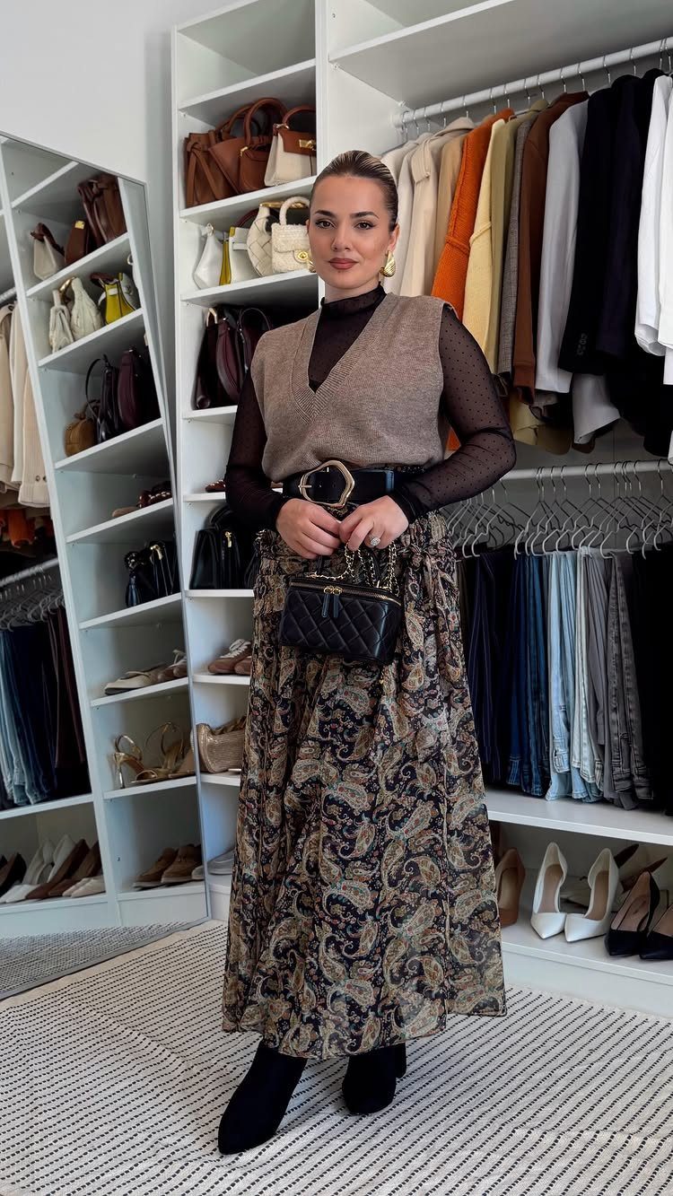 Women's Business Casual: 10 Outfit Ideas for the Office 1 Woman posing in a walk-in closet wearing a patterned midi skirt, sweater vest, and black boots.