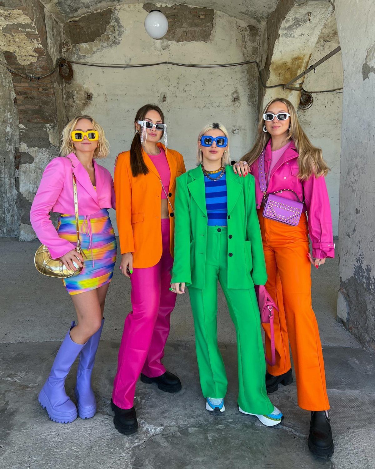 Four women posing in vibrant, brightly colored suits and fashionable outfits against a stone wall.