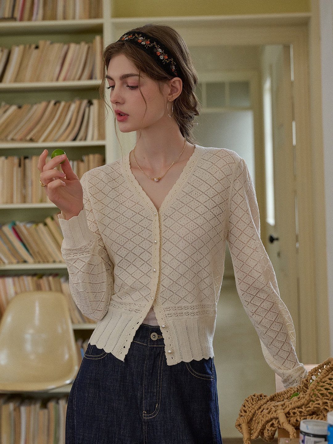 Young woman wearing a cream textured knit cardigan and dark denim jeans in a library setting.