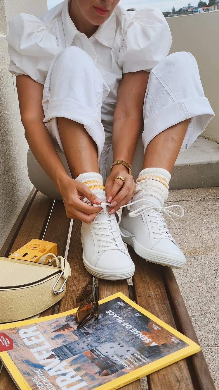 A person sitting on a wooden bench tying their white sneakers and thick socks.