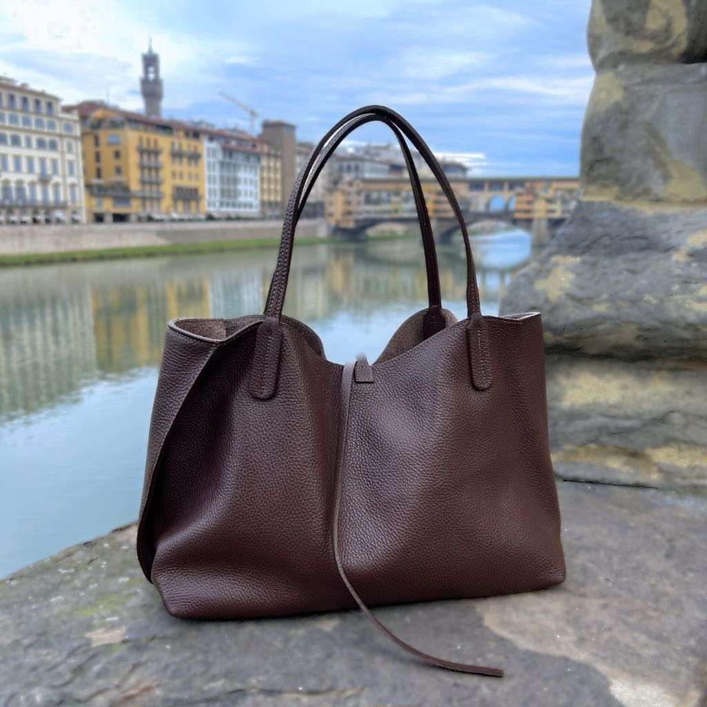 A large dark brown pebbled leather tote bag resting on a stone ledge by a river.