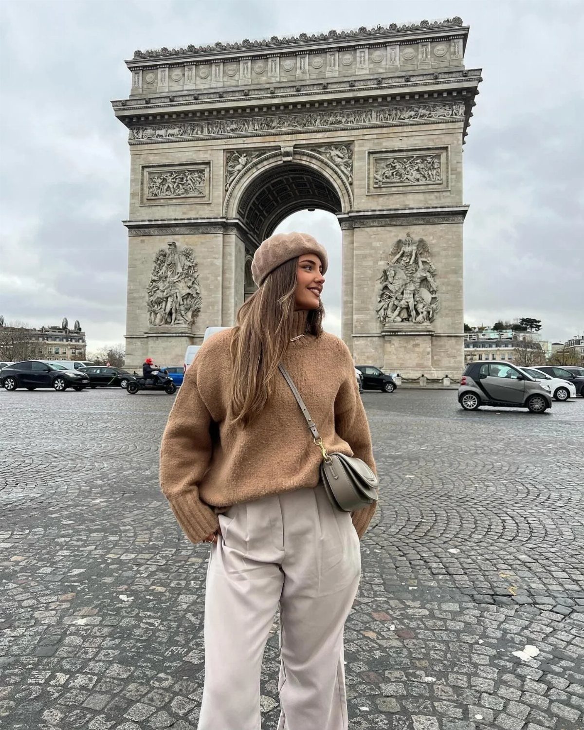 Woman in tan sweater and beige trousers posing in front of the Arc de Triomphe.