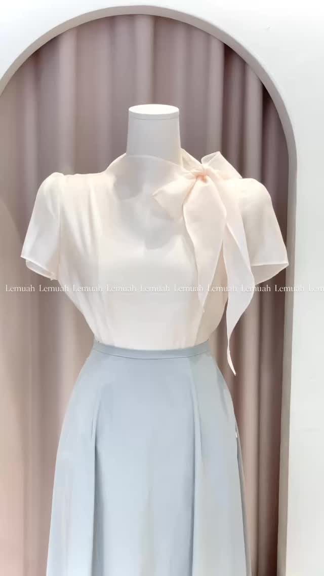 Work Outfits for Women: 7 Smart Office Looks for 2026 7 A mannequin wearing a cream bow-neck blouse and a light blue high-waisted skirt against pink curtains.