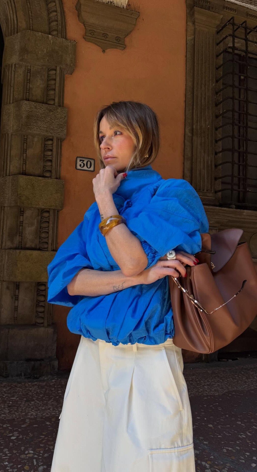 Woman in a blue puffed blouse and white skirt carrying a brown leather handbag.