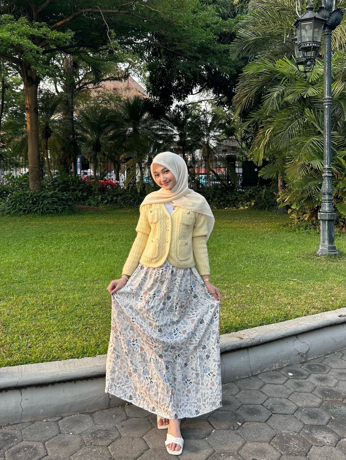 Woman in yellow knit cardigan and floral maxi skirt posing outdoors in a park.