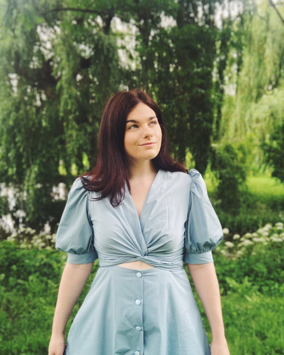 Woman in a light blue puffed-sleeve dress standing before green willow trees.
